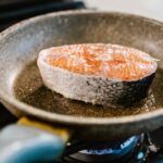 A salmon steak is searing in an iron frying pan on a stove, showcasing cooking detail.