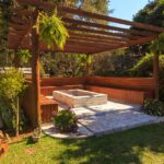 Spacious wooden gazebo in a lush garden setting with green foliage and stone flooring.