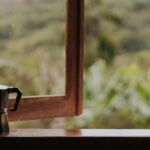 A coffee maker and mug on a window sill, overlooking a lush greenery outdoors.