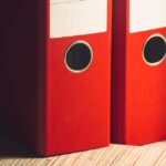 Close-up of two red lever arch files on a wooden desk in a modern office setting.