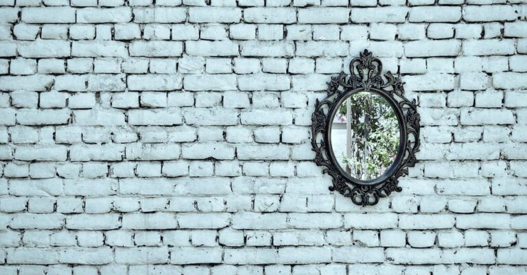 A vintage ornate mirror hanging on a white brick wall reflecting greenery in Kolkata.