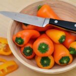 A wooden bowl filled with vibrant orange bell peppers and a knife on a light wooden surface.