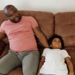 A father and son lounging comfortably on a brown sofa in a cozy indoor setting.