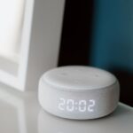 Close-up of a smart speaker with an illuminated 20:02 LED clock display on a desk.