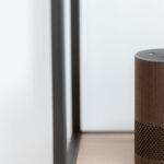 A sleek, modern smart speaker displayed on a wooden shelf in a minimalist room setting.