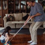 A playful family moment as a father and daughter mop the living room floor together, enjoying quality time.