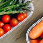 Close-up of fresh asparagus, cherry tomatoes, and carrots in plastic containers on a wooden table.