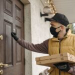A delivery man wearing a mask knocks on a door holding pizzas, symbolizing contactless delivery during the pandemic.