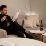 Asian man sitting on a couch reading documents, with a wine bottle on the table.