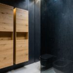 Wooden cupboards hanging on black wall near toilet bowl and shower cabin in contemporary bathroom