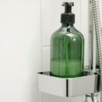 Green dispensers with body lotion on shelf on shower system in bathroom against tiled wall