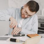 Man wearing glasses using a screwdriver to fix a device on a kitchen table.