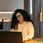 Woman in a yellow sweatshirt using a laptop at home, focusing on remote work in a cozy, modern interior.