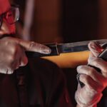 A chef sharpening a knife with precision in a warm-lit kitchen, wearing latex gloves and glasses.