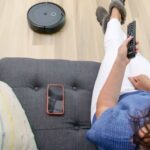 Person relaxing on a sofa controlling a robot vacuum cleaner with a remote.