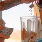 Man adding ingredients to a blender in a van, preparing a fresh smoothie on a sunny day.