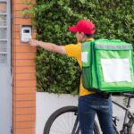 Courier in yellow shirt and red cap delivering a package on a bicycle, pressing a doorbell outdoors.