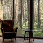 Serene living room with armchairs and lamp, overlooking a lush forest through large windows.